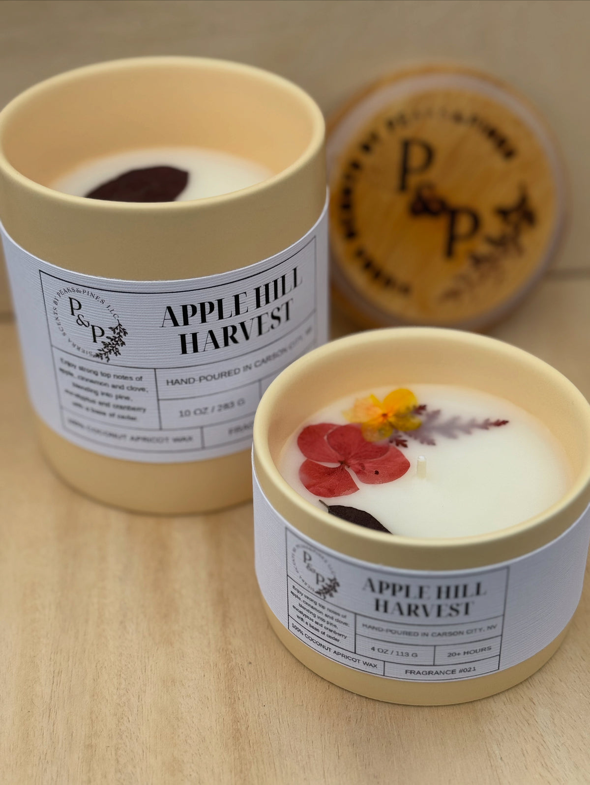 Apple Hill Harvest 10 oz and 4 oz cream ceramic candles showing pressed flowers and leaves in the wax. Branded wood lid in the background. 