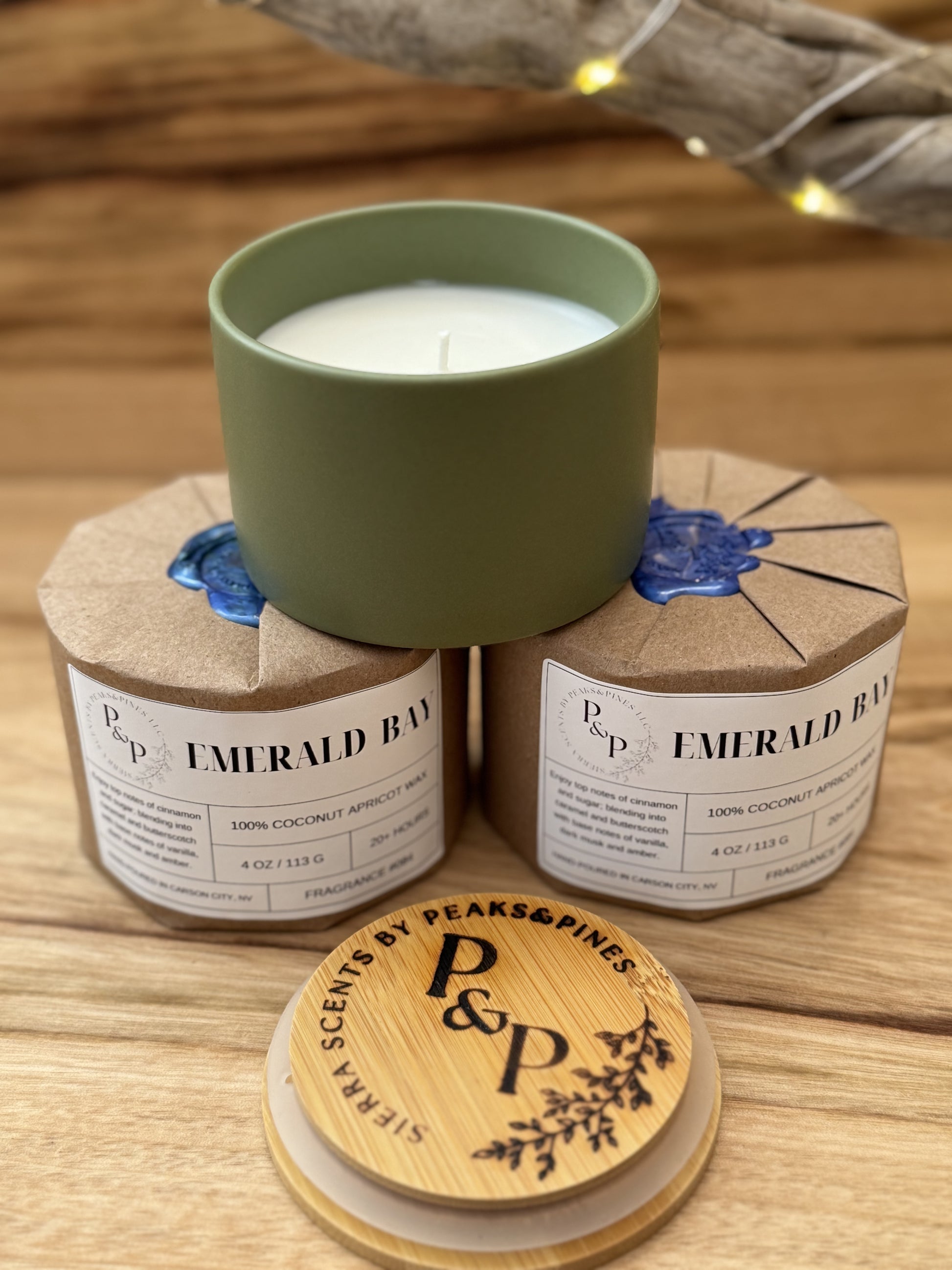 Three Emerald Bay 4 oz candles on a wooden surface, two wrapped in kraft paper with wax seal. One showing the green ceramic and branded wood lid.