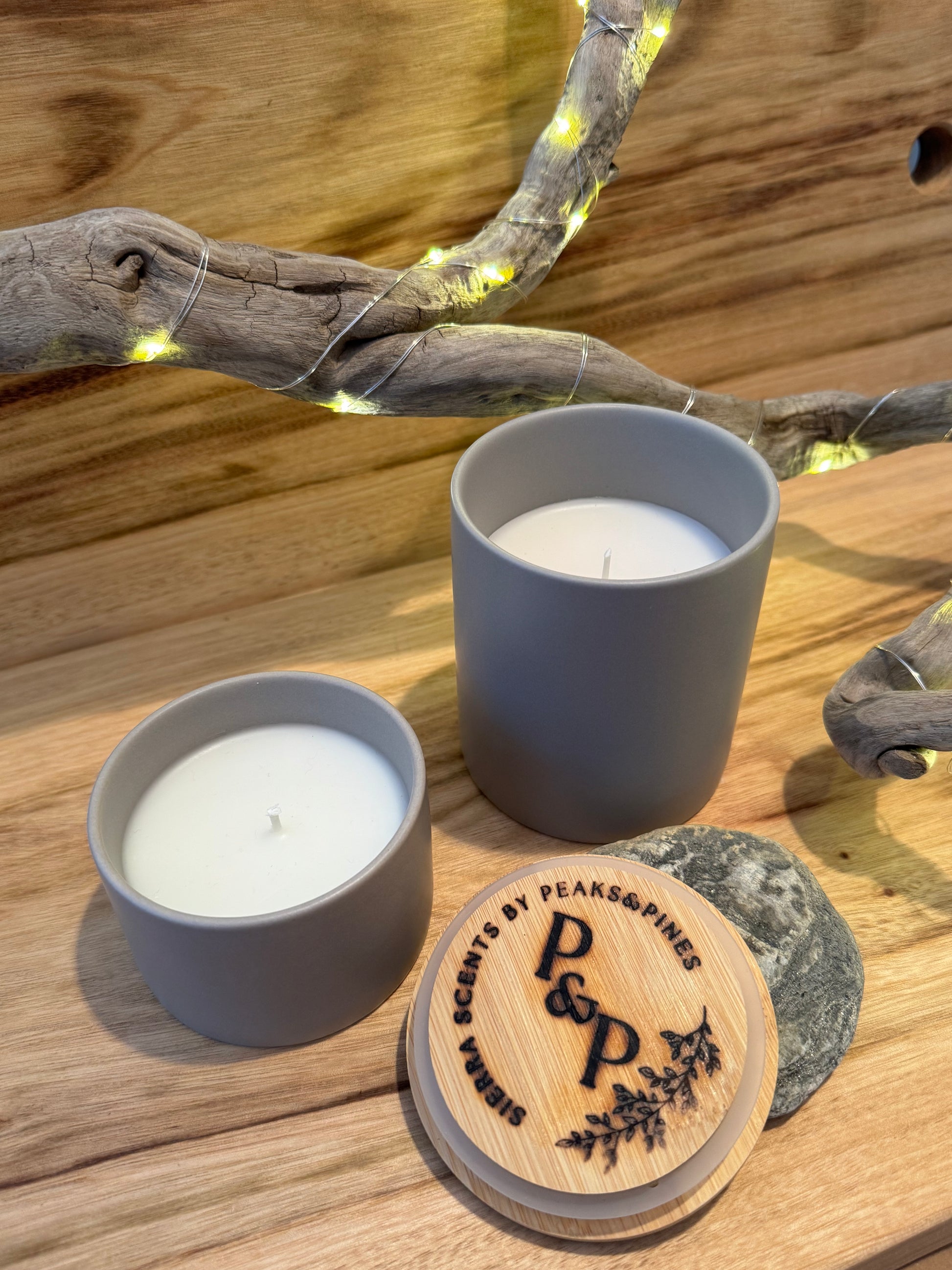 Two gray sand harbor candles with no label on a wooden surface with a branded lid. 