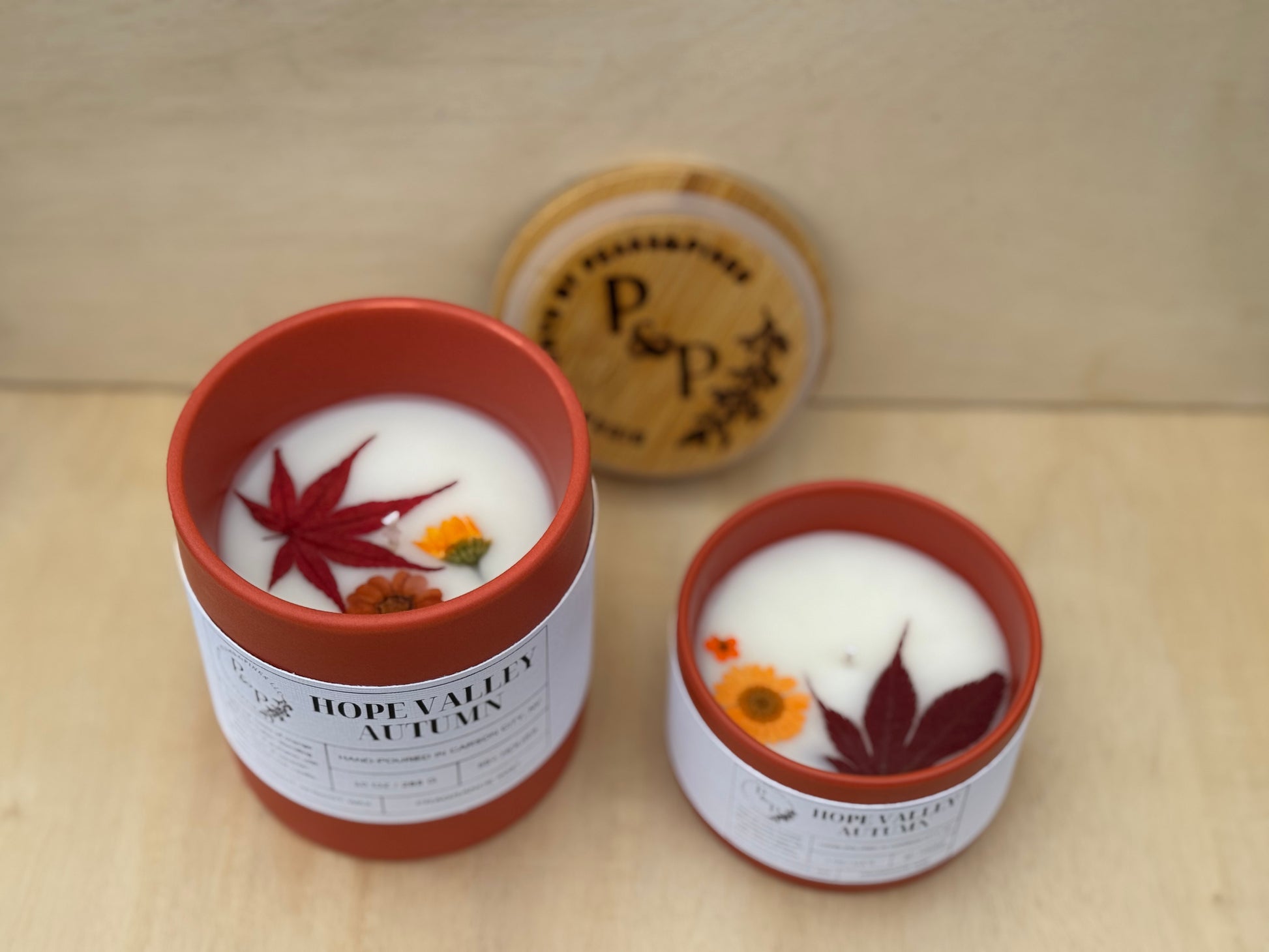 Hope Valley Autumn 4 oz and 10 oz ceramic candles displayed together, both with orange ceramic vessels, pressed leaves and flowers in the wax and white labels with a branded wood lid in the background. 