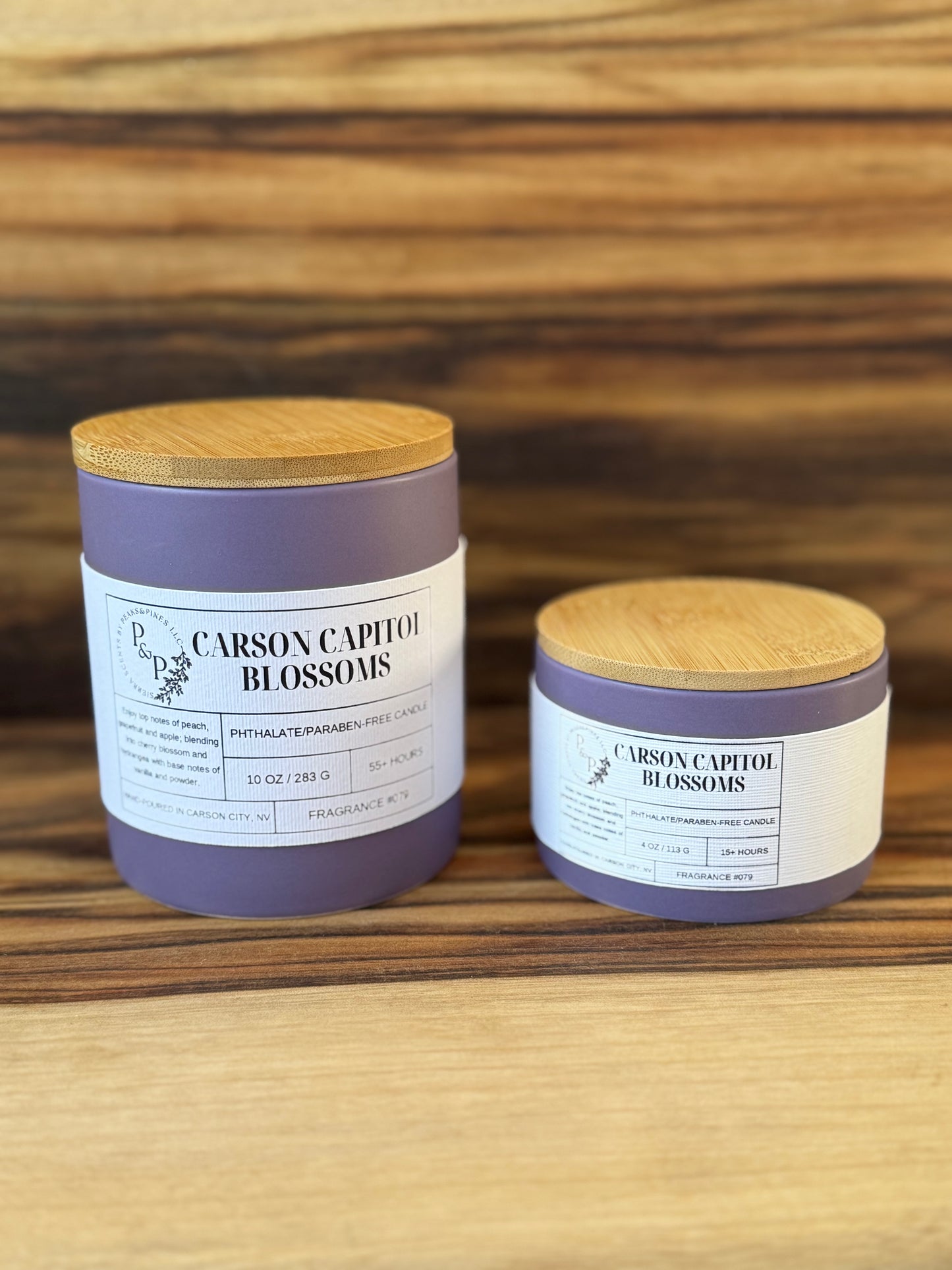 Carson Capitol Blossoms 10 oz and 4 oz purple ceramic candles from the Peaks and Pines displayed together with wood lids and white labels. 
