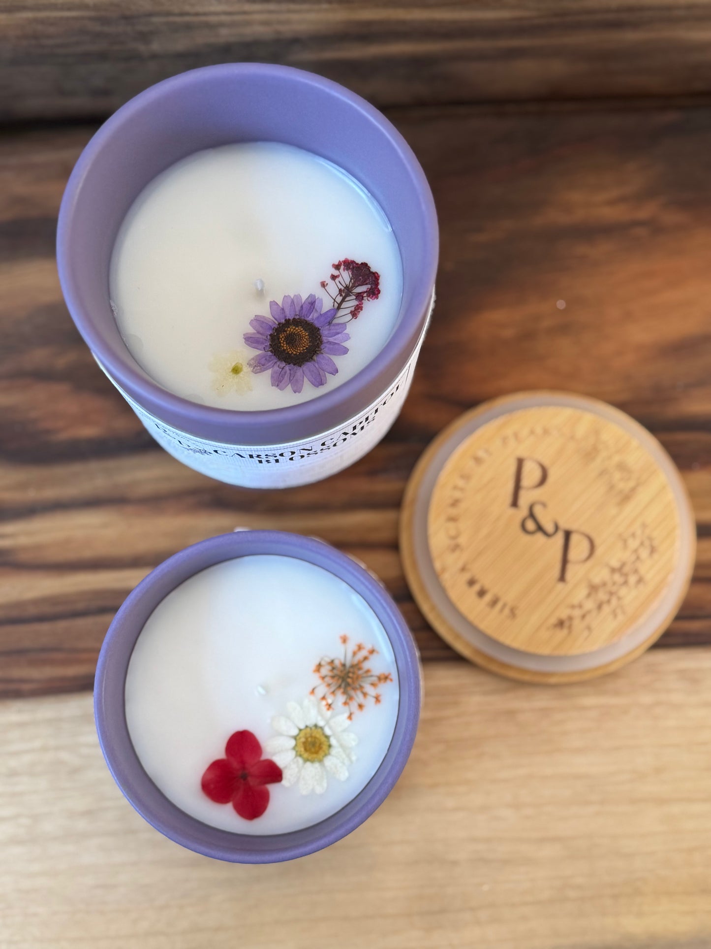 Carson Capitol Blossoms 10 oz and 4 oz purple ceramic candles from the Peaks and Pines botanical impressions line displayed together. Showing pressed flowers in the wax. 