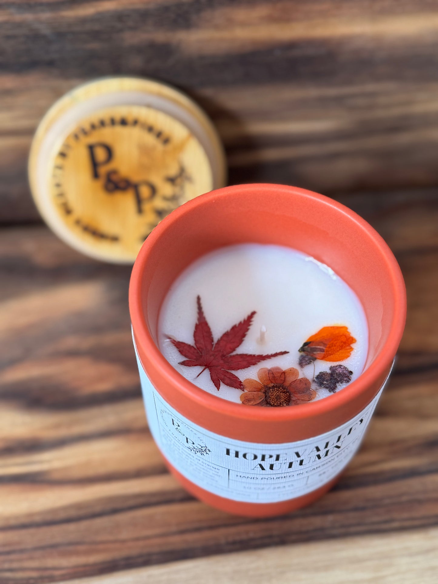 Hope Valley Autumn 10 oz orange ceramic candle by Peaks and Pines.  Pressed flowers and leaves are shown in the wax of the candle with the branded wood lid in the back. 

