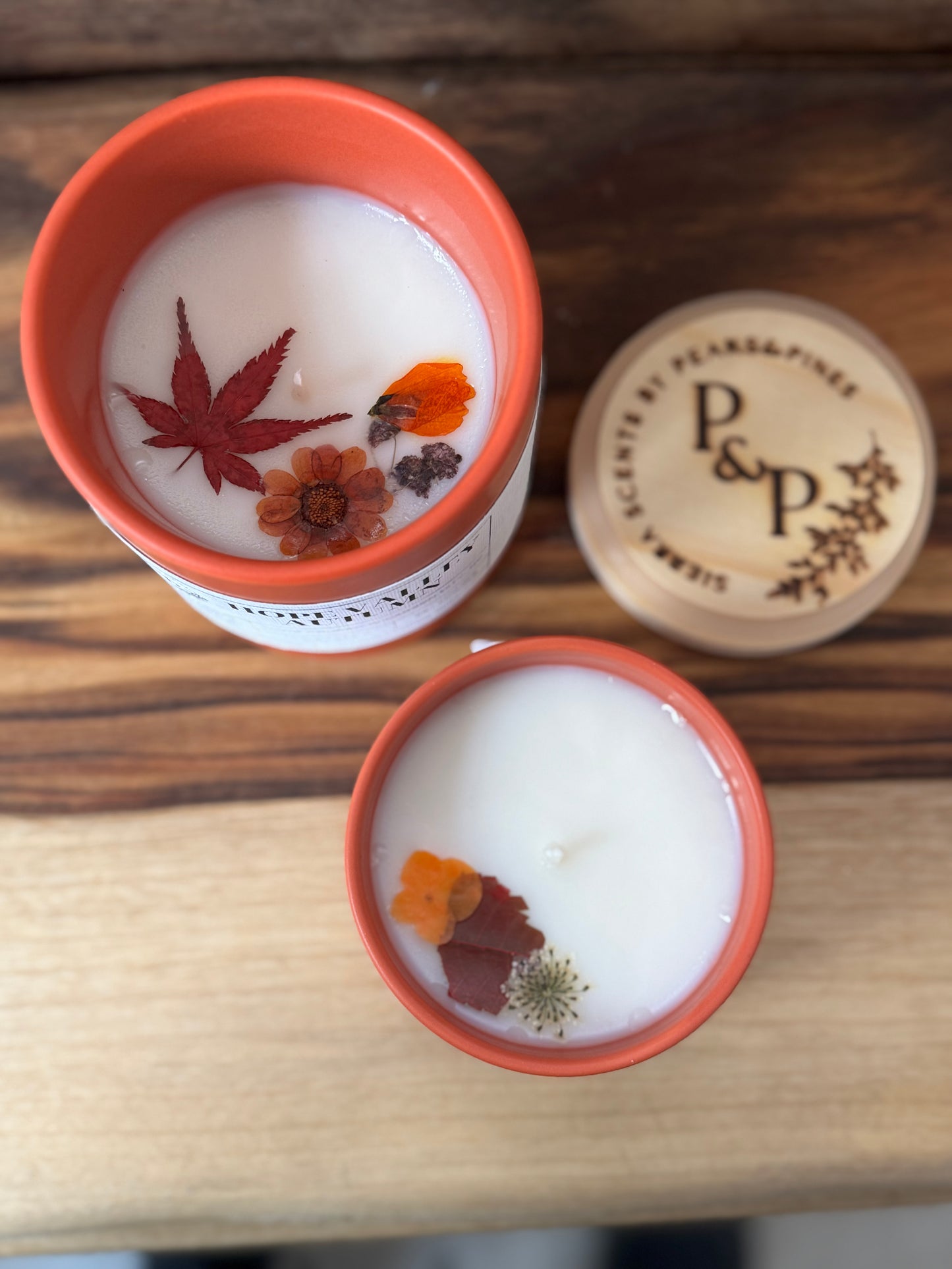 Two  Hope Valley Autumn candles with pressed leaves and flowers in the wax in orange vessels on a wooden surface, with a branded wood lid labeled 'P&P' in the background.