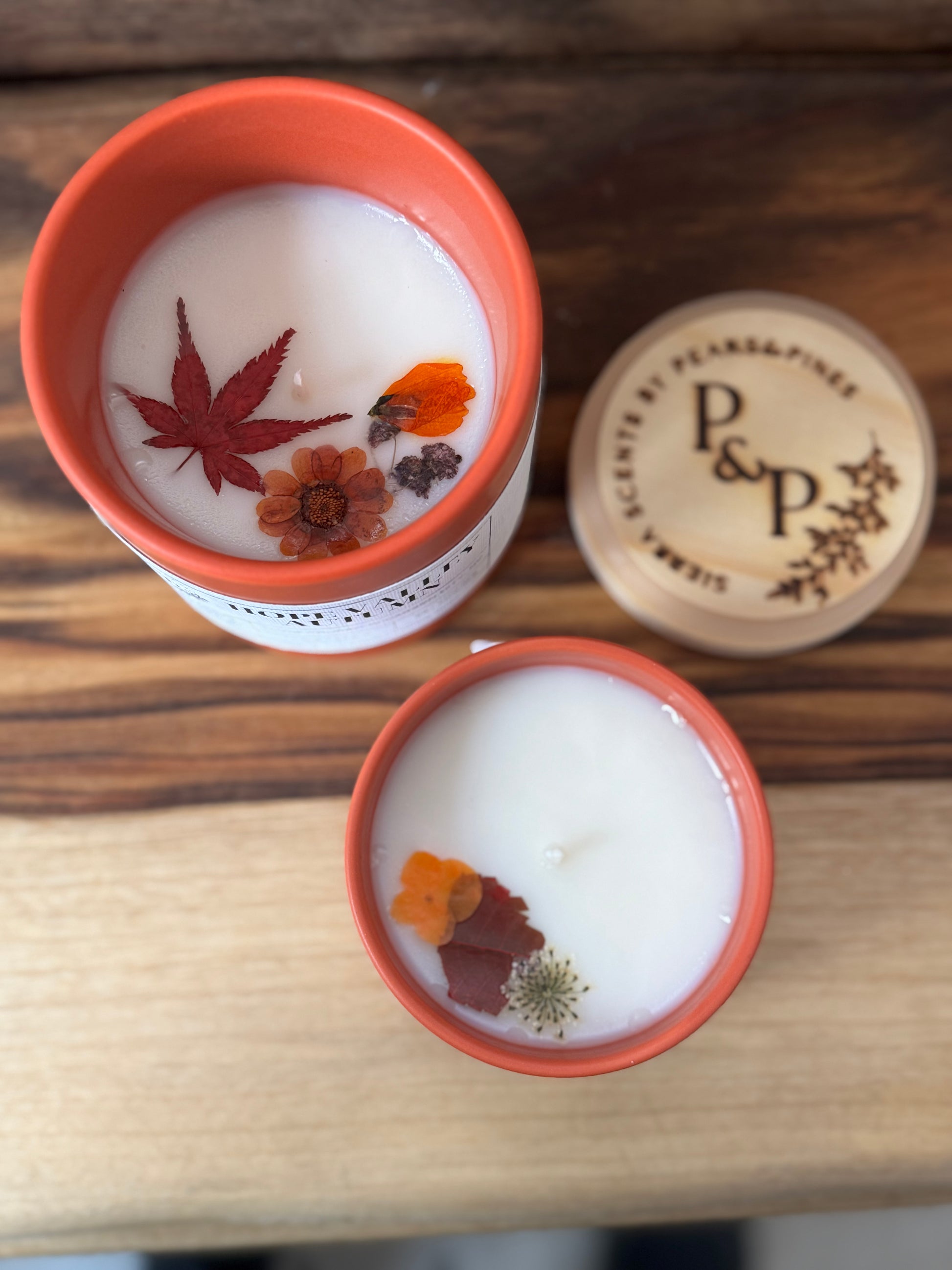 Two  Hope Valley Autumn candles with pressed leaves and flowers in the wax in orange vessels on a wooden surface, with a branded wood lid labeled 'P&P' in the background.